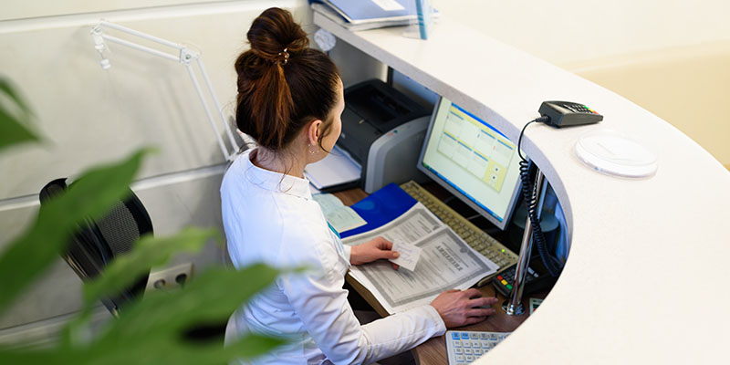 A woman working at a reception desk in an office setting with a computer monitor and keyboard visible.