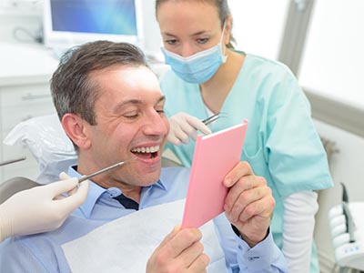 The image shows a man in a dental office, smiling and holding up a pink card with the word 'Congratulations' written on it. He is seated in a chair with medical equipment around him, wearing a surgical mask, and there's a female dental professional in the background assisting him.